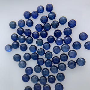 以下が含まれることがあります： 白い表面に散らばる、丸く磨かれた深い青色の宝石のコレクション。宝石は滑らかで反射性の高い表面を持ち、わずかに色合いが異なり、光と影の微妙な遊びを生み出しています。全体的な効果は、エレガンスと自然の美しさです。