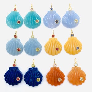 May include: A collection of twelve shell-shaped pendants arranged in three rows. Each pendant is crafted from colored stone with a gold-tone bail and two small, round, bezel-set gemstones in various colors embedded on the surface of the shell.