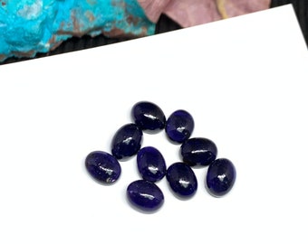 May include: A group of 10 oval-shaped, dark blue gemstones. The gemstones are arranged in a loose, scattered pattern on a white surface.
