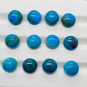 May include: Twelve round, blue and green cabochon gemstones with gold flecks. The gemstones are arranged in three rows of four on a white surface.