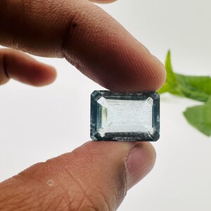 May include: A light blue gemstone with a rectangular emerald cut. The gemstone is held between two fingers.