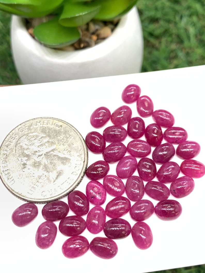 Puede incluir: Una colecci&oacute;n de aproximadamente 30 piedras preciosas de rub&iacute; rosa de forma ovalada. Las piedras preciosas est&aacute;n esparcidas sobre una superficie blanca, con una moneda de veinticinco centavos de d&oacute;lar estadounidense para la escala.
