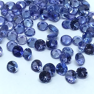 May include: A collection of round, faceted, blue gemstones. The gemstones are scattered on a white surface.