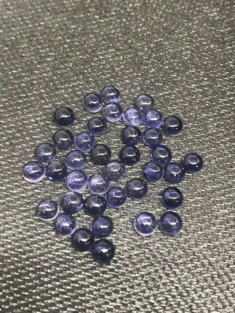 May include: A collection of round, purple gemstones, likely amethyst, displayed on a textured gray surface.