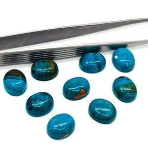 May include: A collection of polished, oval-shaped gemstones in shades of teal and turquoise, with brown and gold veining. A pair of silver tweezers is positioned above the stones. The gemstones are displayed against a white background.