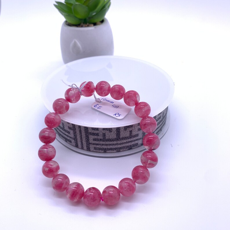 May include: Pink gemstone bracelet with round beads. The bracelet is on a white display stand with a black and white patterned ribbon. The tag on the bracelet reads "10mm 1A R3 EE".