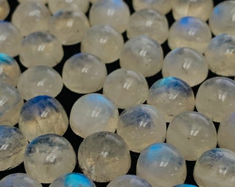 Puede incluir: Una colección de cabujones redondos de piedra lunar blanca con destellos azules iridiscentes. Las piedras son lisas y pulidas, y están dispuestas en un patrón aleatorio.