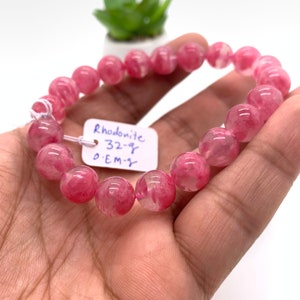 May include: A pink Rhodonite gemstone bracelet with a tag that reads "Rhodonite 32-g 0.EM-g".
