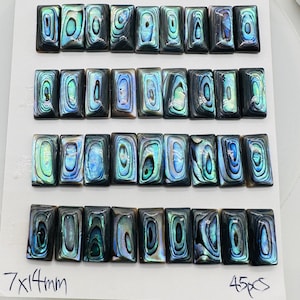 May include: A collection of 45 iridescent abalone shell cabochons, each measuring 7x14mm, arranged in rows on a white background. The cabochons have a blue, green, and black color pattern.