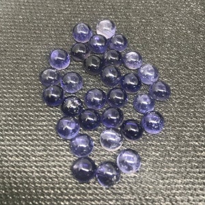 May include: A group of round, purple gemstones with a slight shimmer. The gemstones are arranged in a loose pile on a textured gray surface.