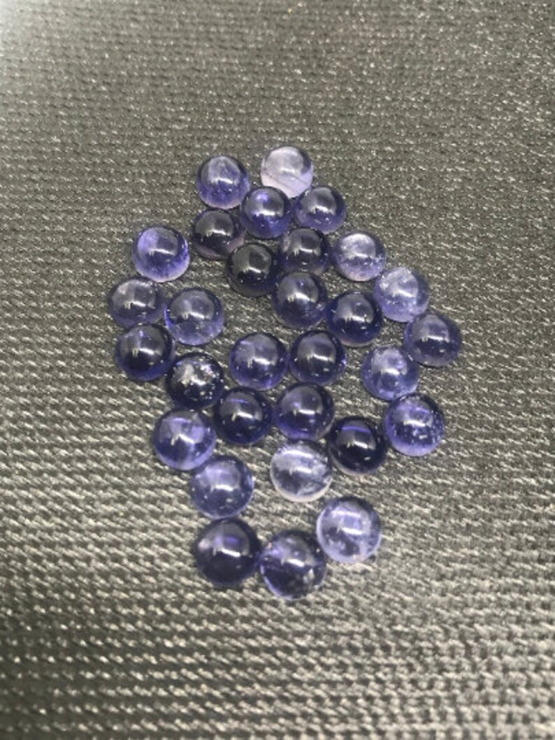May include: A group of round, purple gemstones with a slight shimmer. The gemstones are arranged in a loose pile on a textured gray surface.