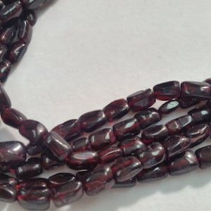 May include: A strand of dark red gemstone beads, likely garnet, with a smooth, polished finish. The beads are oval-shaped and have a slightly irregular, natural look.