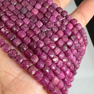 May include: A strand of pink ruby beads, each bead is faceted and has a square shape. The beads are strung on a red thread.