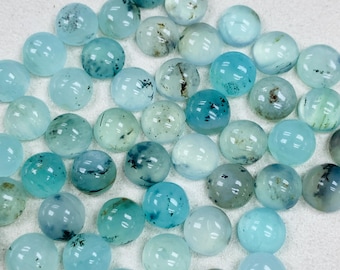 May include: A collection of round, polished gemstones in varying shades of blue and teal. Some stones have dark inclusions, creating unique patterns. The gemstones are scattered on a white surface, showcasing their translucent quality.