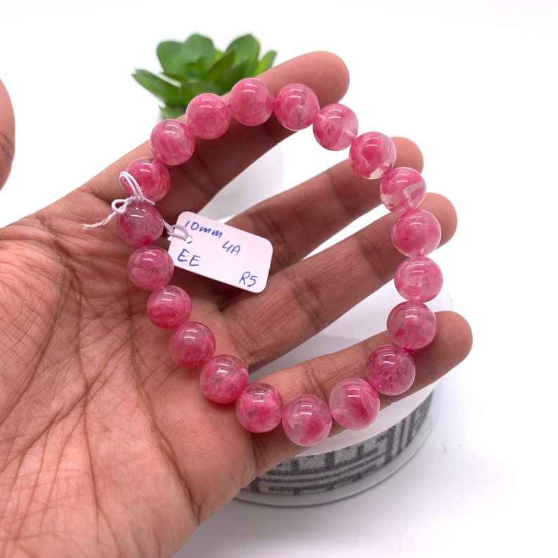 May include: A pink and white gemstone bracelet with round beads. The bracelet is 10mm wide and has a tag that says "10mm UA EE R5".