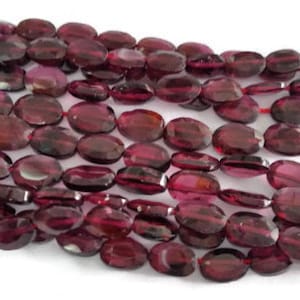 May include: A strand of oval-shaped, faceted, red garnet beads. The beads are smooth and polished, with a deep red colour.