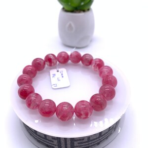 May include: Pink gemstone bracelet with round beads. The bracelet is on a white stand with a black and white pattern. The tag on the bracelet reads "12mm, Lip, MX, P2".