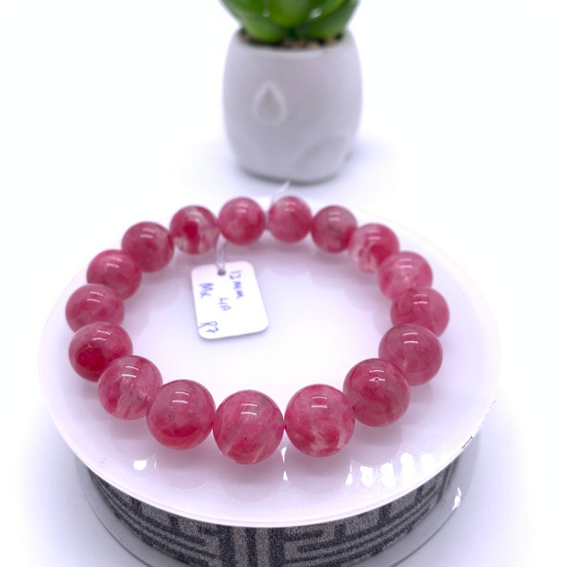 May include: Pink gemstone bracelet with round beads. The bracelet is on a white stand with a black and white pattern. The tag on the bracelet reads "12mm, Lip, MX, P2".