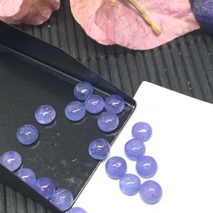 May include: A collection of round, light purple gemstones, some in a black tray and others on a white surface.