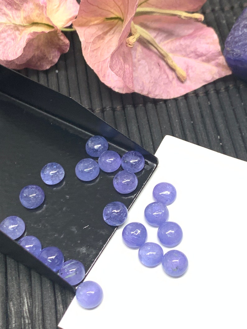 May include: A collection of round, light purple gemstones, some in a black tray and others on a white surface.