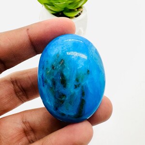 May include: A smooth, oval-shaped blue gemstone with green and white marbling. The gemstone is being held in a hand.