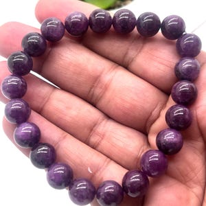 May include: A stretch bracelet composed of numerous smooth, round, deep purple beads. The beads exhibit natural variations in color intensity and texture, creating a consistent circular pattern. The bracelet is designed to fit comfortably around a wrist.