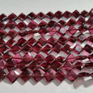 May include: A strand of dark red gemstone beads, each bead is faceted and shaped like a square with beveled edges.
