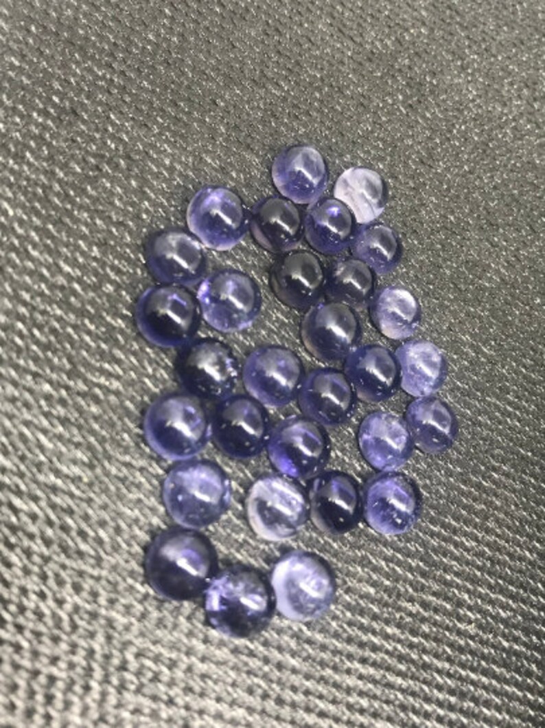 May include: A collection of round, purple gemstones with a smooth, polished finish. The stones are arranged in a loose cluster on a gray textured surface.