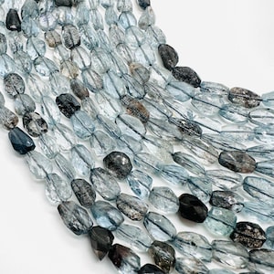 May include: A strand of light blue and gray faceted gemstone beads. The beads are oval shaped and have a translucent appearance. The beads are strung on a black thread.