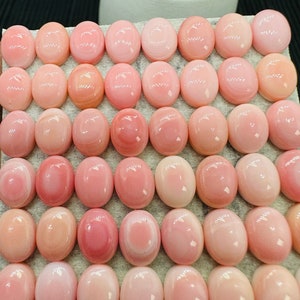 Peut inclure: Une collection de 48 cabochons ovales en coquille rose et blanche. Les cabochons sont disposés en rangées sur une surface blanche.