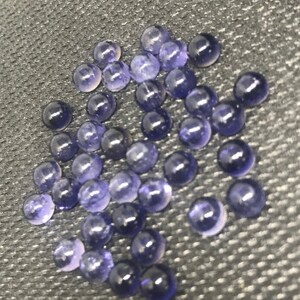 May include: A collection of small, round, purple gemstones. The gemstones are smooth and shiny, and they are scattered on a dark gray surface.
