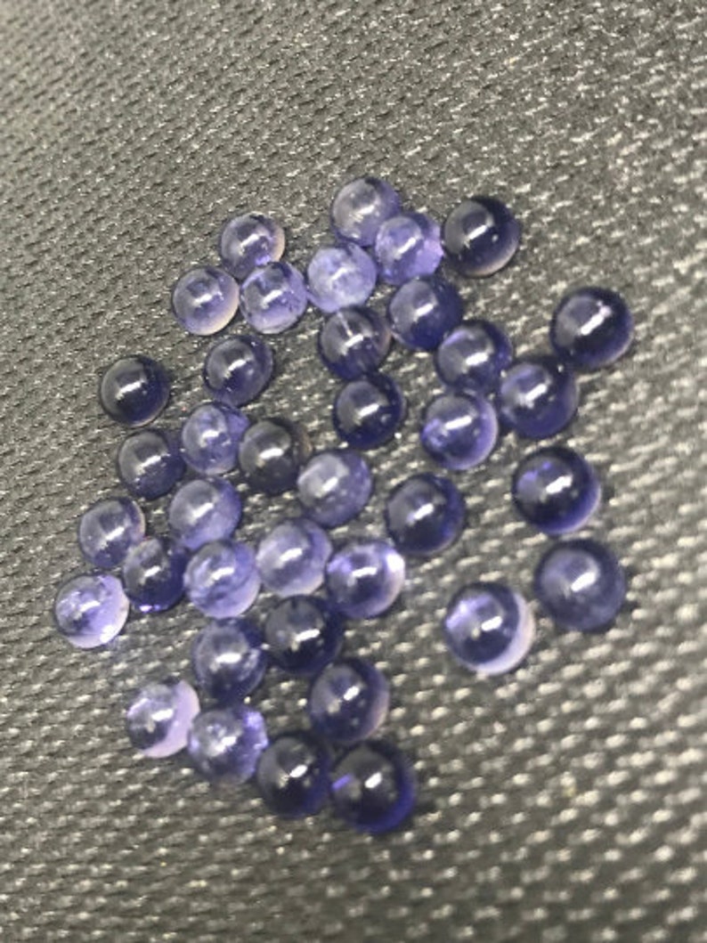 May include: A collection of small, round, purple gemstones. The gemstones are smooth and shiny, and they are scattered on a dark gray surface.