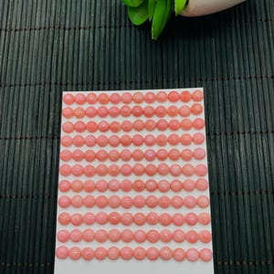 May include: A collection of 132 pink cabochon gemstones, each approximately 6mm in diameter, arranged on a white card. The gemstones are a soft, uniform pink color. The card also has the number 289 written on it.