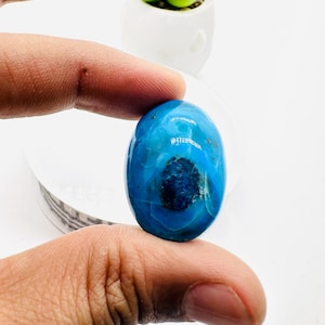 May include: A bright blue oval cabochon gemstone with a dark blue center and white veins. The gemstone is being held by a hand.