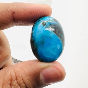 May include: A smooth, oval-shaped cabochon gemstone with a blue and black marbled pattern. The stone is held in a hand.