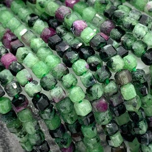 Może przedstawiać: Szereg zielonych, czarnych i różowych koralików z kamieni szlachetnych. Koraliki są fasetowane i mają kwadratowy kształt. Koraliki są nawleczone na przezroczystą nić.