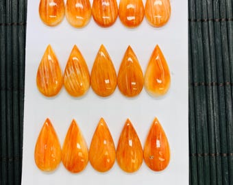 Könnte beinhalten: Fünfzehn orangefarbene, tropfenförmige Edelsteine, angeordnet auf einer weißen Oberfläche. Jeder Edelstein hat eine polierte, glänzende Oberfläche mit natürlichen Maserungen. Die Edelsteine messen 1,2 cm x 1,4 cm. Die Gesamtpalette ist warm, mit Variationen von Orange und Creme.