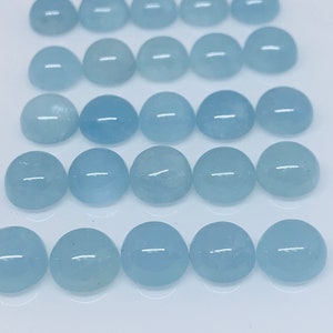 May include: Twenty-four light blue, cabochon-cut gemstones arranged in rows on a white surface. The stones have a smooth, rounded top and a slightly translucent appearance. These gemstones are likely aquamarine, a popular choice for jewelry.