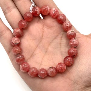 May include: A beaded bracelet featuring round, translucent, pink-red beads. The beads have a marbled appearance with lighter pink and white inclusions. The bracelet is strung on a white elastic cord and is displayed on a hand.