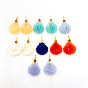 Puede incluir: Una colección de doce colgantes con forma de concha dispuestos en tres filas. Cada colgante presenta una concha de piedra preciosa tallada con una anilla de metal dorado y un pequeño acento de piedra preciosa engastada en varios colores.