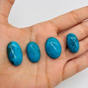 May include: Four polished, oval-shaped gemstones in varying sizes, displayed on a hand. The stones are a vibrant turquoise color with darker blue and green veining. The gemstones have a smooth, glossy surface, reflecting light.