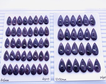 May include: Two sets of teardrop-shaped, polished gemstones in shades of purple and white. The gemstones are arranged on white cards, with one set labeled "8x16mm" and the other "10x20mm". The cards are placed on a white tray.