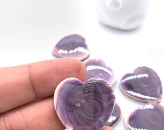 May include: Heart-shaped polished shells in shades of purple and white. The shells have a smooth, glossy finish and a natural, swirling pattern. Several shells are visible, with one held in a hand. The shells are likely used for jewelry or craft projects.