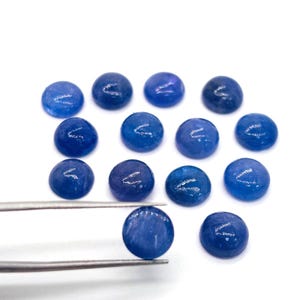 May include: A collection of round, polished blue gemstones. One gemstone is held by silver tweezers, showcasing its smooth, reflective surface. The other gemstones are scattered on a white background, displaying variations in color and texture.