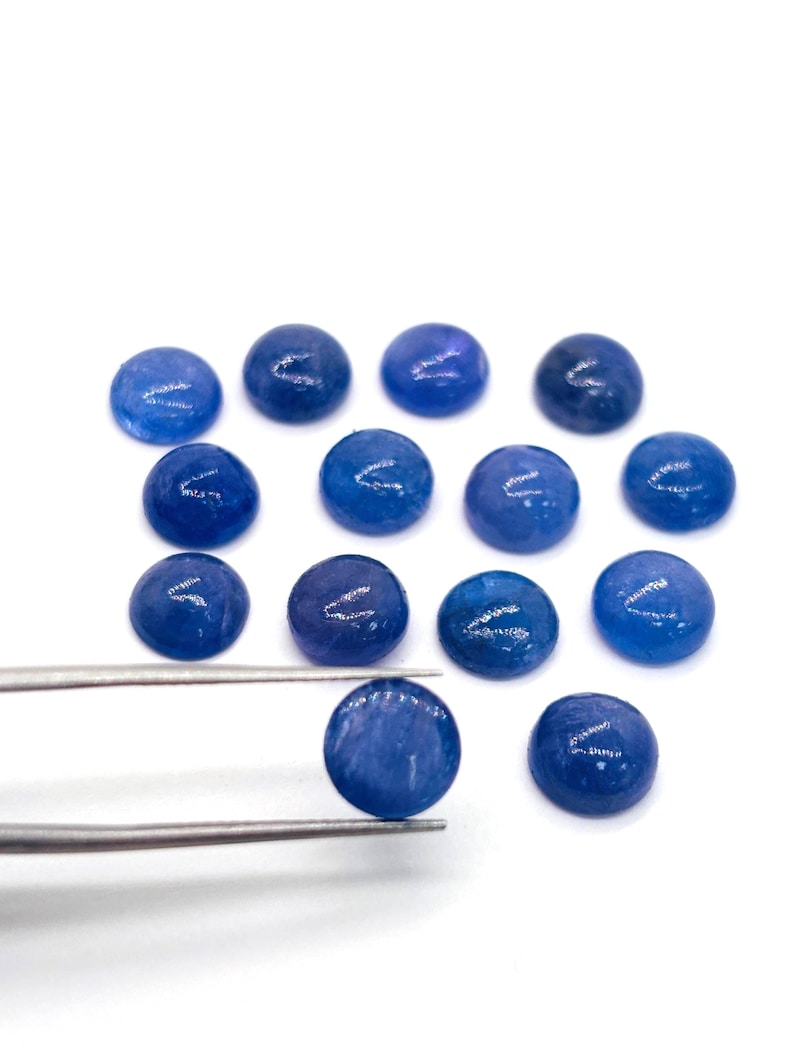 May include: A collection of round, polished blue gemstones. One gemstone is held by silver tweezers, showcasing its smooth, reflective surface. The other gemstones are scattered on a white background, displaying variations in color and texture.