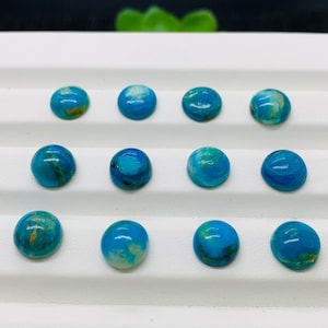 May include: Twelve round, blue and white cabochon gemstones with gold flecks, arranged in three rows of four on a white surface.