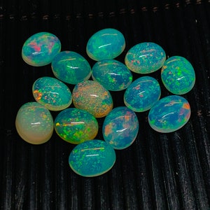May include: A group of 12 oval-shaped opal gemstones with a variety of green, blue, and yellow iridescent colors. The stones are arranged in a cluster on a black background.