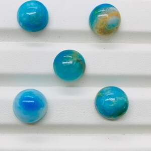 May include: Five round, blue and white cabochon gemstones with a smooth, polished finish. The gemstones are arranged in a row on a white surface.