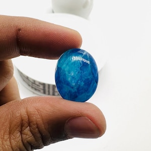 May include: A blue agate cabochon with white swirls and a glossy finish. The cabochon is oval shaped and is being held by a hand.