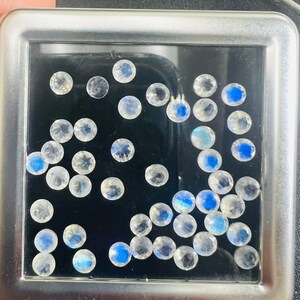 May include: A close-up of a metal box containing a collection of round, iridescent white gemstones. The gemstones are scattered randomly on a black background.